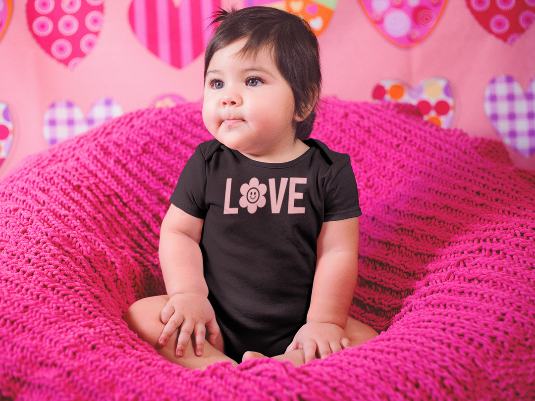 A baby in a pink blanket sits comfortably, showcasing the Love Onesie from Worlds Worst Tees. This infant fine jersey bodysuit is 100% cotton, featuring ribbed knitting for durability and easy plastic snaps for changing.