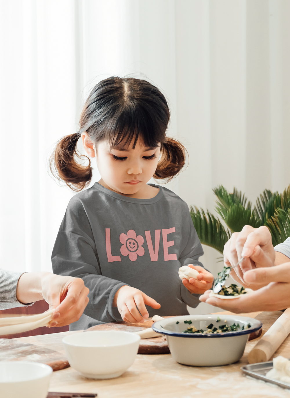 A toddler long-sleeve tee featuring a girl with ponytails and a woman holding a spoon with food, a girl holding a piece of dough, and a girl holding clay in her hands. Made of 100% combed ringspun cotton fine jersey for durability and comfort.