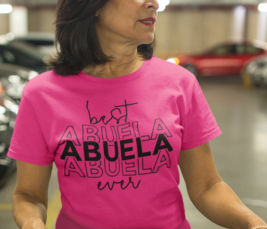 A woman in a pink shirt pushing a shopping cart, showcasing the Best Abuela Ever Tee. 100% ring-spun cotton, garment-dyed for coziness. Relaxed fit, durable double-needle stitching, no side-seams for a tubular shape.