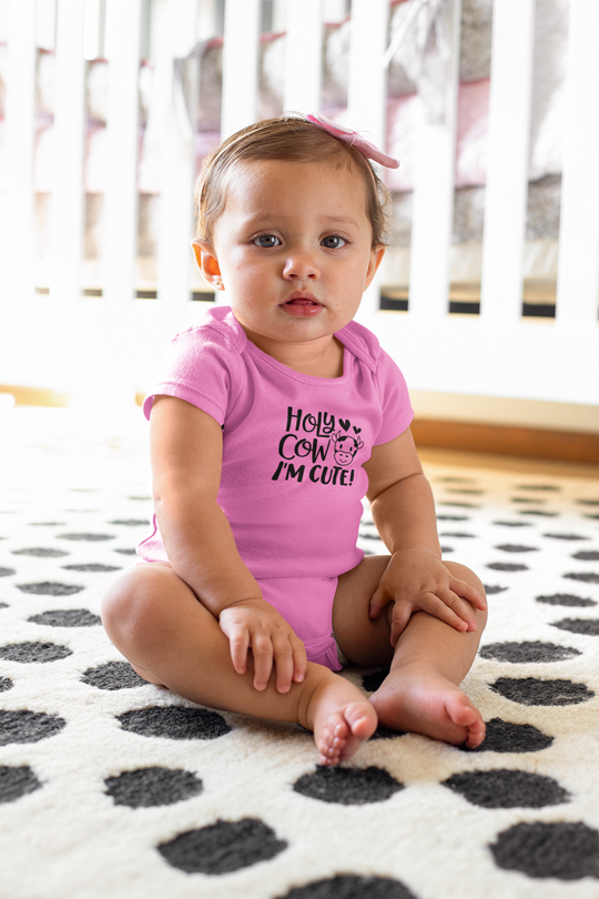 A baby in a pink shirt sits on the floor, showcasing the Holy Cow I'm Cute Onesie by Worlds Worst Tees. This infant bodysuit is 100% cotton, featuring ribbed bindings and plastic snaps for easy changing access.
