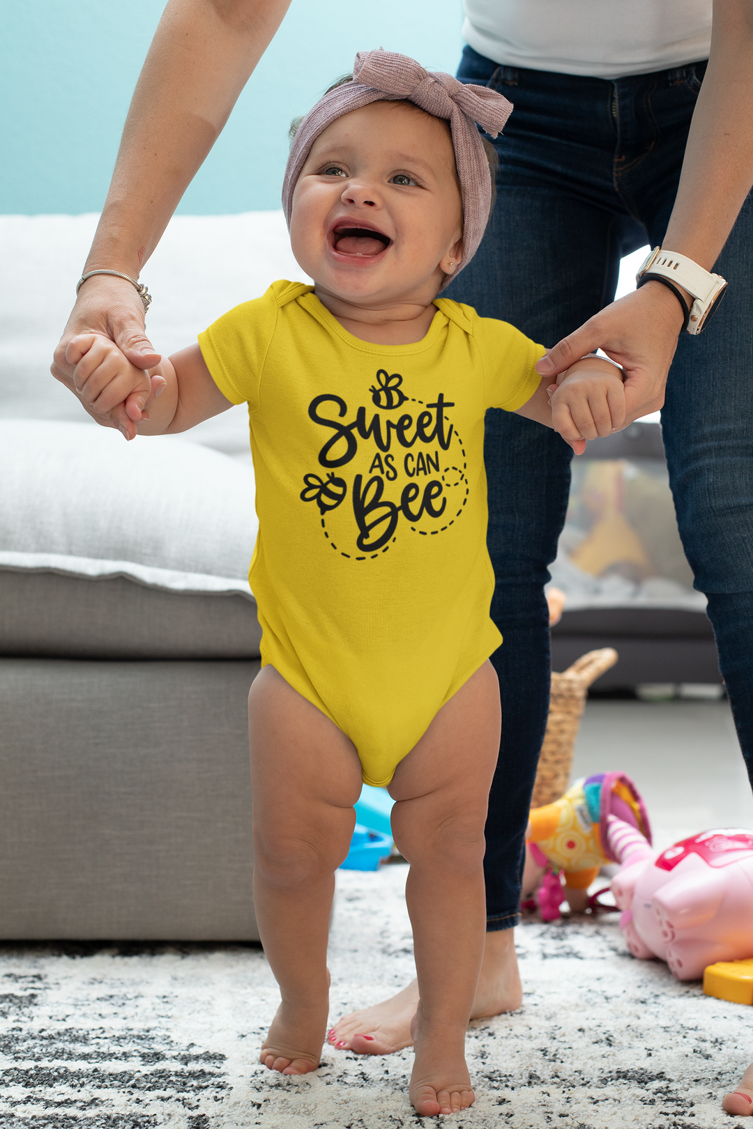 A baby in a yellow Sweet as can Be onesie, standing in front of a woman's hands. This infant fine jersey bodysuit is 100% cotton, with ribbed knitting for durability and plastic snaps for easy changing access.