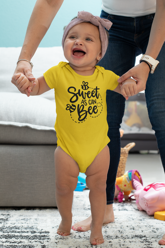 A baby in a yellow Sweet as can Be onesie, standing in front of a woman's hands. This infant fine jersey bodysuit is 100% cotton, with ribbed knitting for durability and plastic snaps for easy changing access.