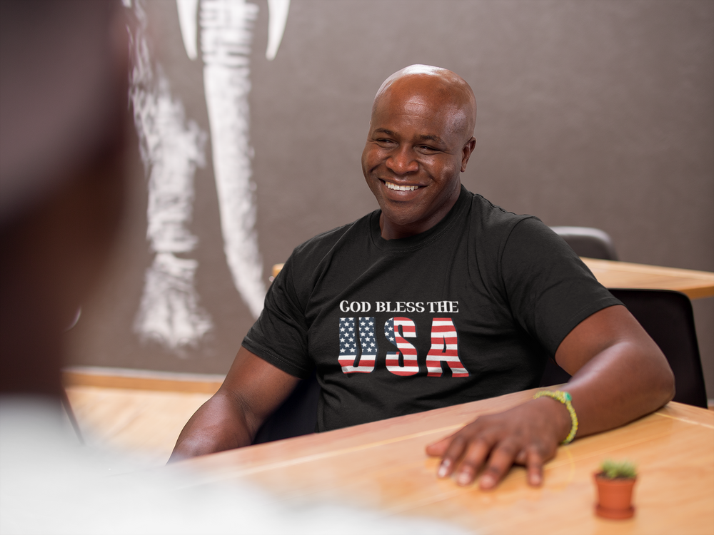A man in a black shirt with a flag, smiling at a table. Unisex God Bless The USA Dudes Tee: 100% cotton, classic fit, sustainably sourced materials. From 'Worlds Worst Tees'.