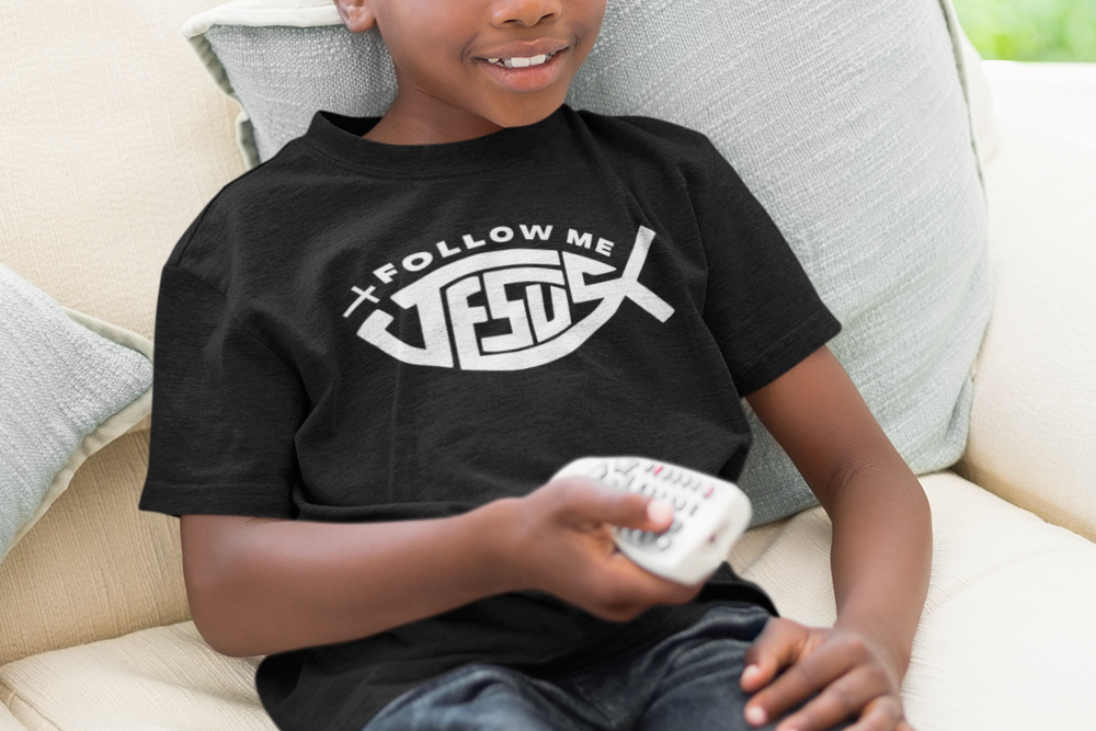 A boy sitting on a couch holding a remote control, wearing a Jesus Follow Me Kids Tee. Comfortable 100% cotton, classic fit, ideal for everyday wear. Visible features: tee, remote control, couch.