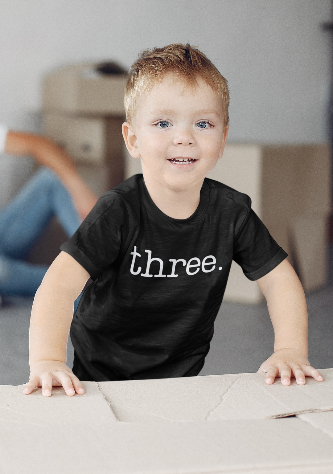 A toddler stands on a table, smiling at the camera. Three Toddler Tees: classic fit, durable with double-needle collar, sleeve, and bottom hems. Made of 100% Ringspun cotton.