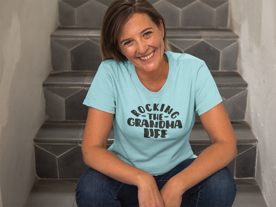 A smiling woman in jeans sits on stairs, embodying the relaxed fit of the Rocking the Grandma Life Tee. Made of 100% ring-spun cotton for coziness and durability.